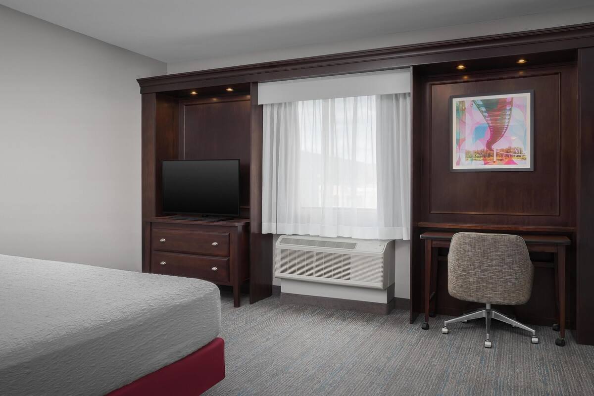 A hotel room with a bed, a desk with a chair, a television, a window with curtains, and a radiator.