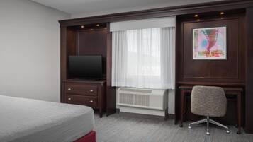 Une chambre d’hôtel comprenant un lit, un bureau avec une chaise, une télévision, une fenêtre avec des rideaux et un radiateur.