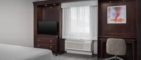 A hotel room with a bed, a desk with a chair, a television, a window with curtains, and a radiator.