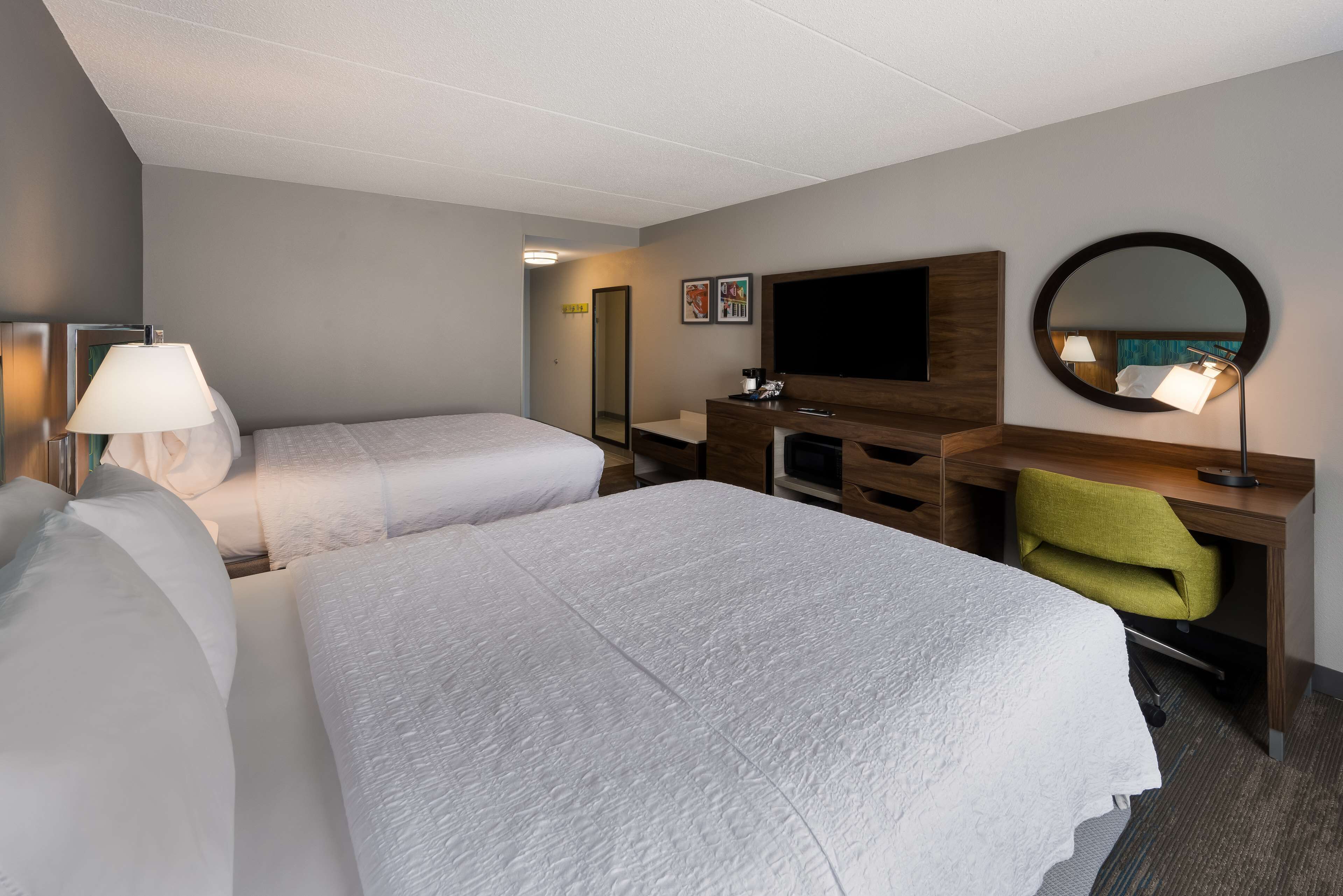 Habitación de hotel con dos camas, un escritorio con silla, televisor, espejo y lámpara.