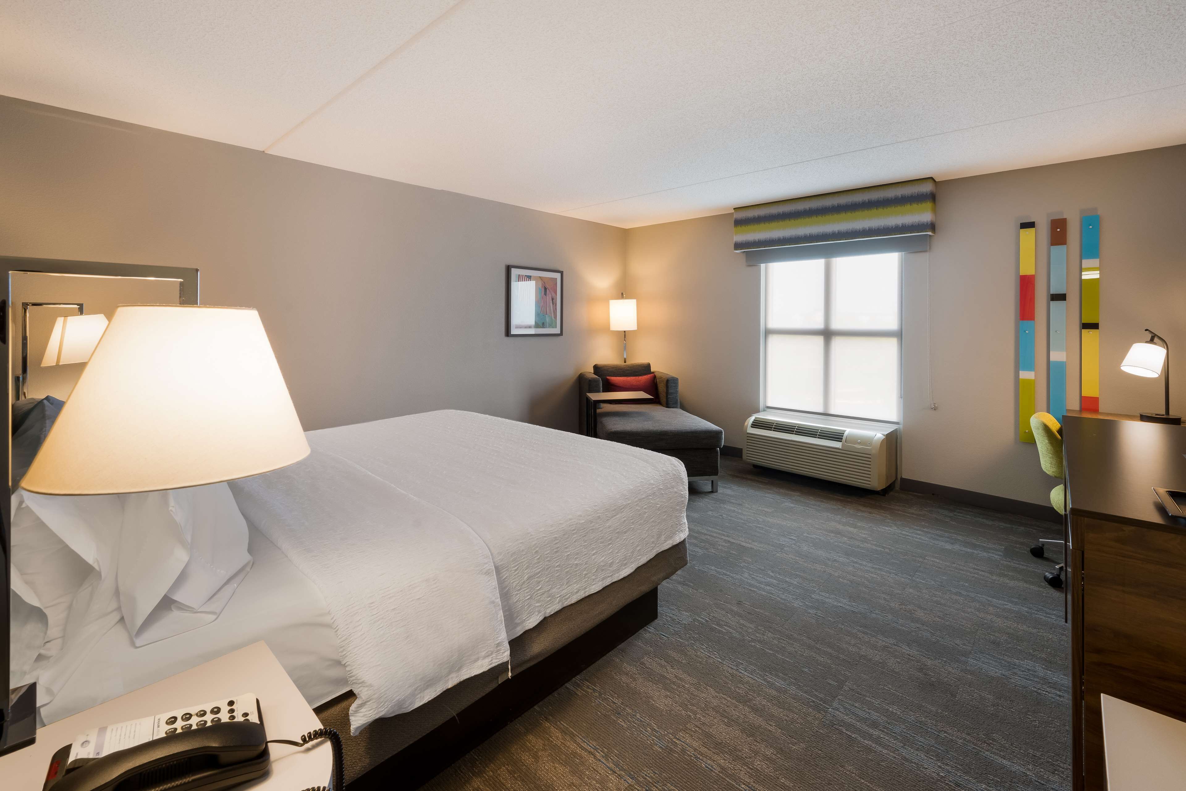 A hotel room with a large bed, bedside lamp, air conditioning unit, and a desk with a lamp.