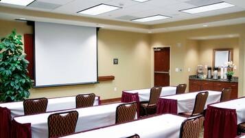 Un salón de conferencias con mesas, sillas, una pantalla de proyector, una planta y una estación de café.