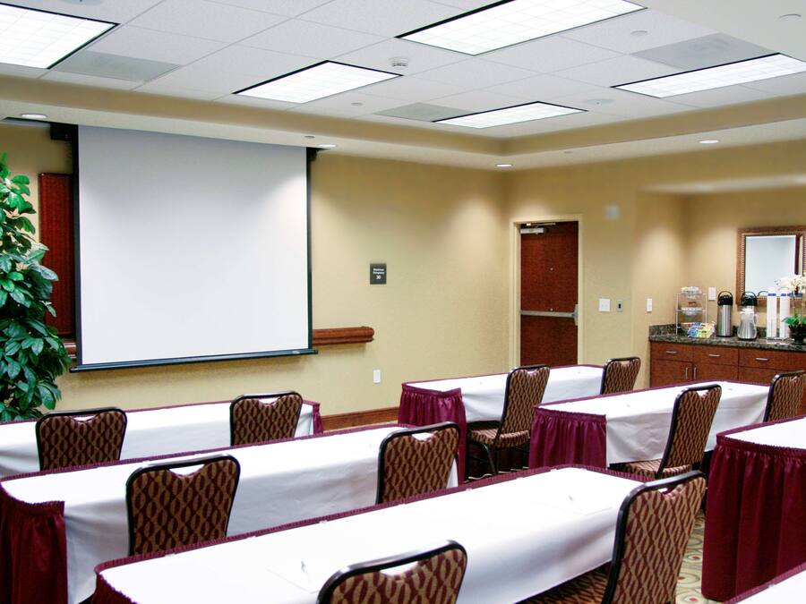 Un salón de conferencias con mesas, sillas, una pantalla de proyector, una planta y una estación de café.