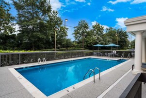 Piscina rectangular al aire libre con agua azul cristalina, rodeada por un muro de piedra bajo y una cerca de metal. Se observan escalones que dan acceso a la piscina, además de algunos sombrillas y sillas al fondo.
