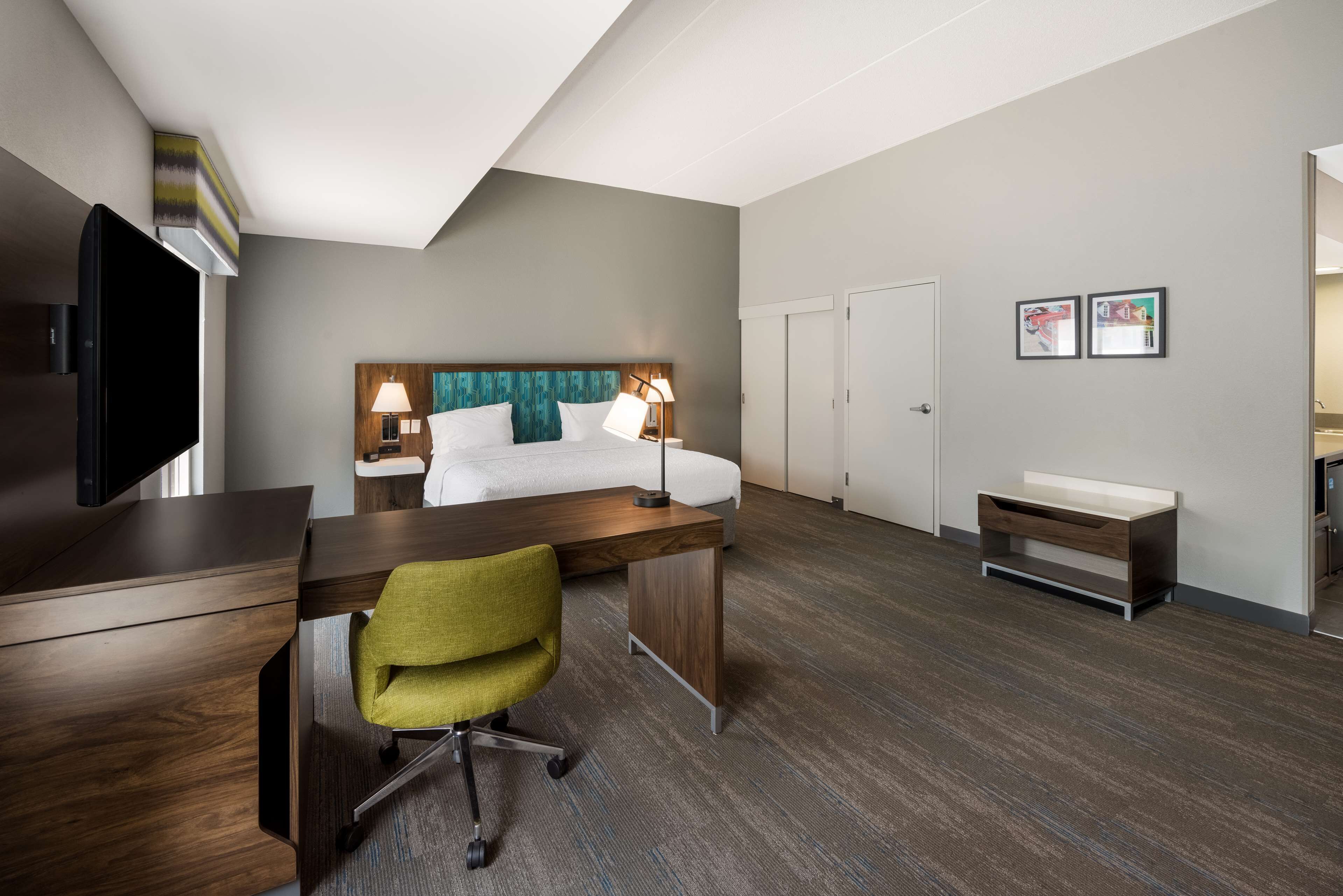 A hotel room with a bed, desk, chair, TV, and artwork on the wall.