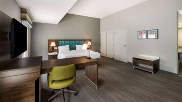 A hotel room with a bed, desk, chair, TV, and artwork on the wall.