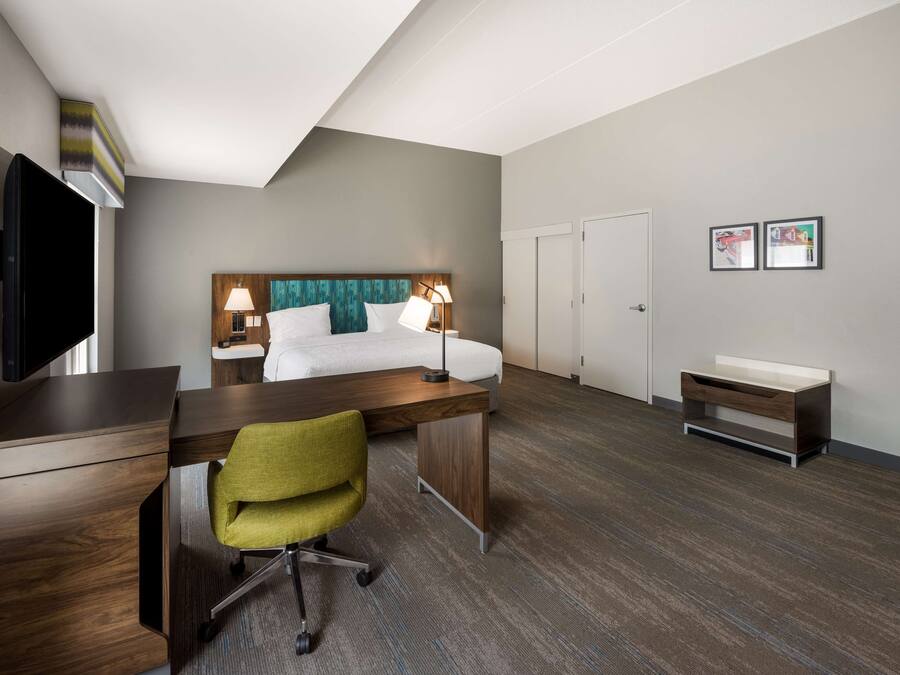 A hotel room with a bed, desk, chair, TV, and artwork on the wall.