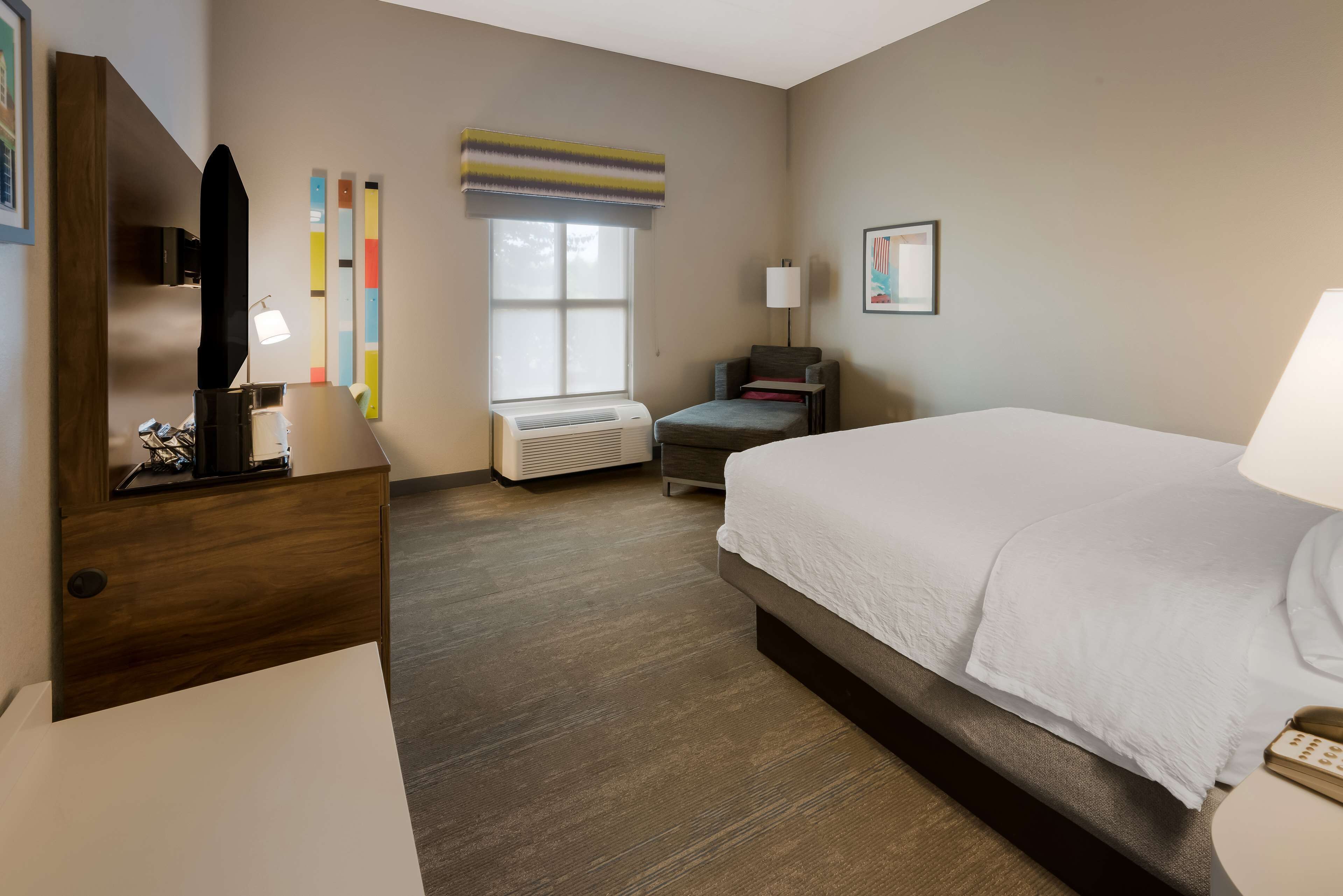 Une chambre d’hôtel équipée d’un lit, d’un bureau, d’une chaise, d’une lampe, d’une fenêtre avec stores et d’un téléviseur fixé au mur.