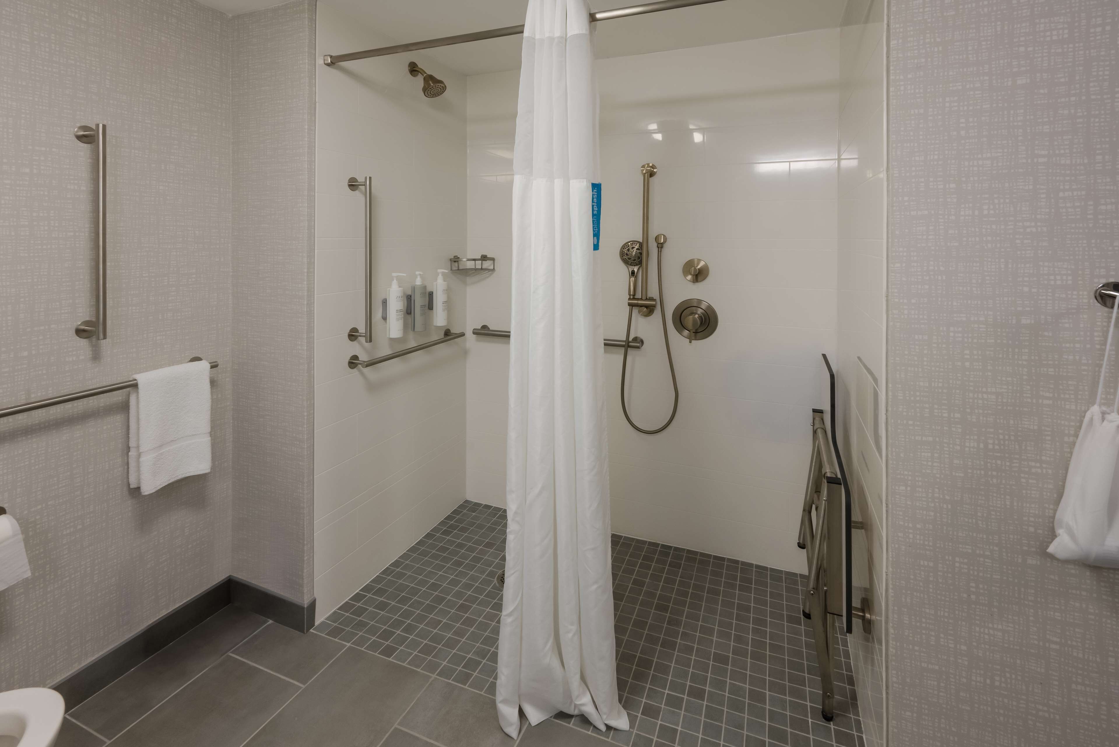 Un baño moderno con ducha a nivel, barras de apoyo, cortina de ducha blanca y suelo de baldosas.