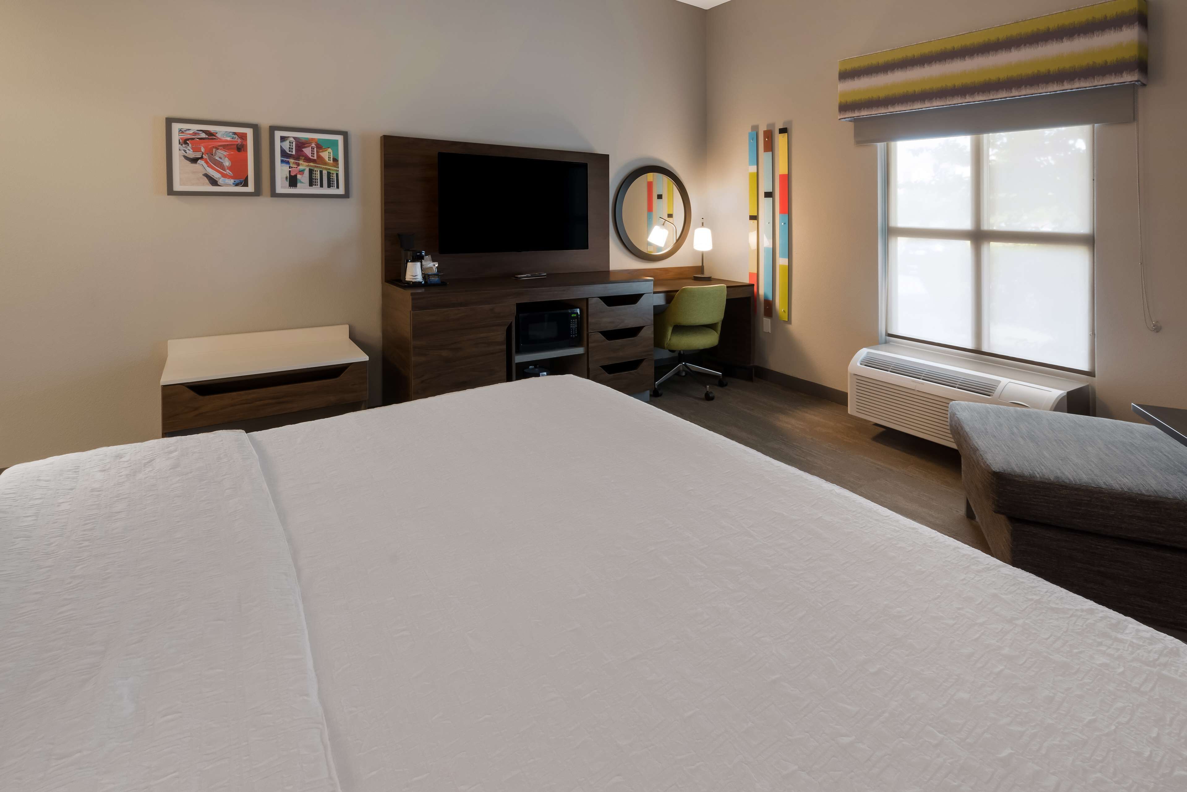 A hotel room with a large bed, a desk with a flat-screen TV, a microwave, a chair, and a window with blinds.