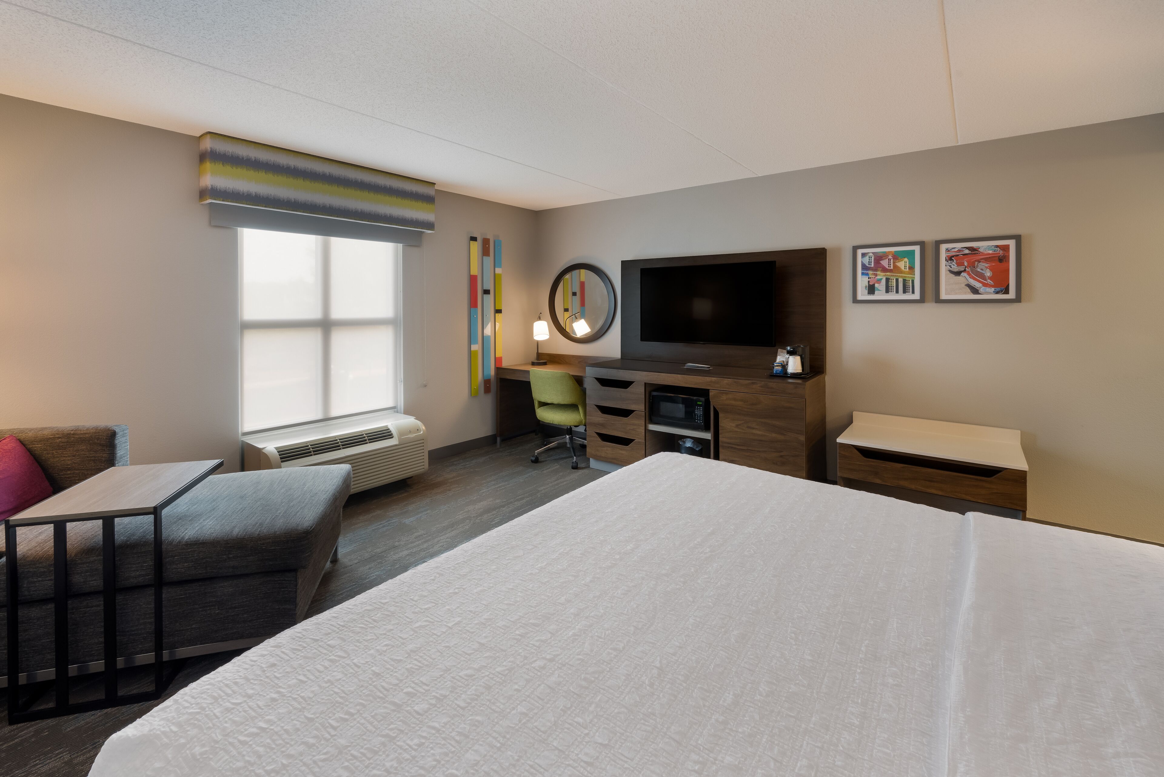 A hotel room with a large bed, a desk with a chair, a flat-screen TV, a small table, and a window with blinds.
