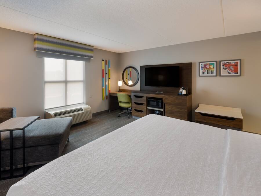 A hotel room with a large bed, a desk with a chair, a flat-screen TV, a small table, and a window with blinds.