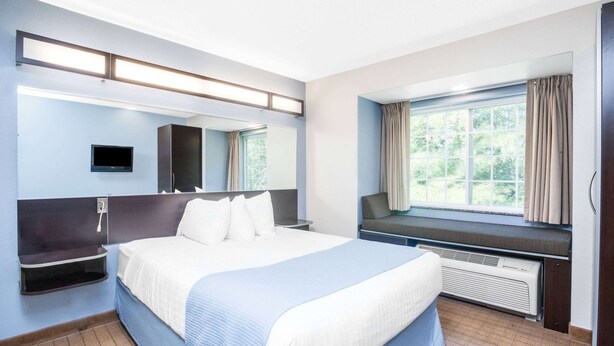 Hotellihuone, jossa on suuri sänky, yöpöytä, televisio, ikkuna verhoilla ja sisäänrakennettu penkki.