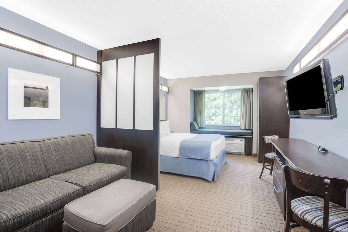A hotel room with a sofa, a bed, a desk with a chair, a television, and a window with curtains.