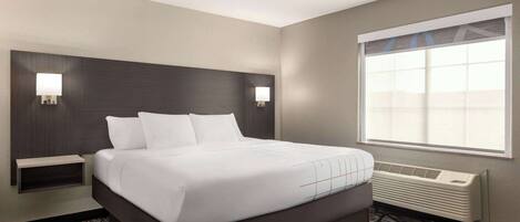 A hotel room with a large bed, two bedside tables, a window with blinds, a wall-mounted heater, and a patterned carpet.