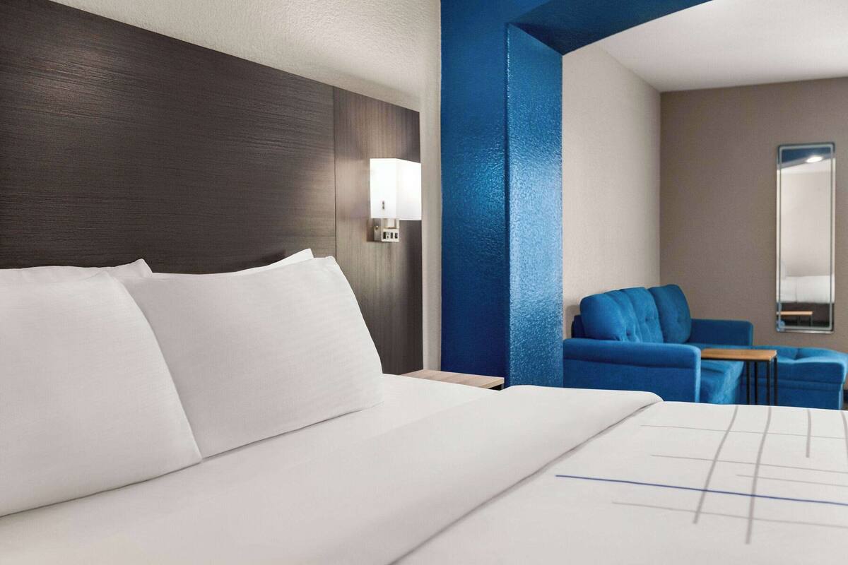Une chambre d’hôtel avec un grand lit, une table de chevet, un fauteuil bleu, une petite table et une lampe fixée au mur.