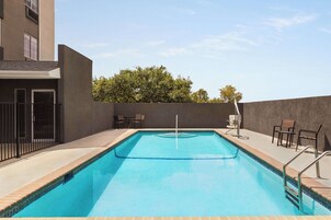Ein rechteckiger Swimmingpool mit gefliestem Rand, umgeben von einer niedrigen Ziegelmauer und einem Metallgeländer. Auf der rechten Seite stehen zwei Stühle und ein Tisch, auf der linken Seite eine Tür mit einem Metallgitter.