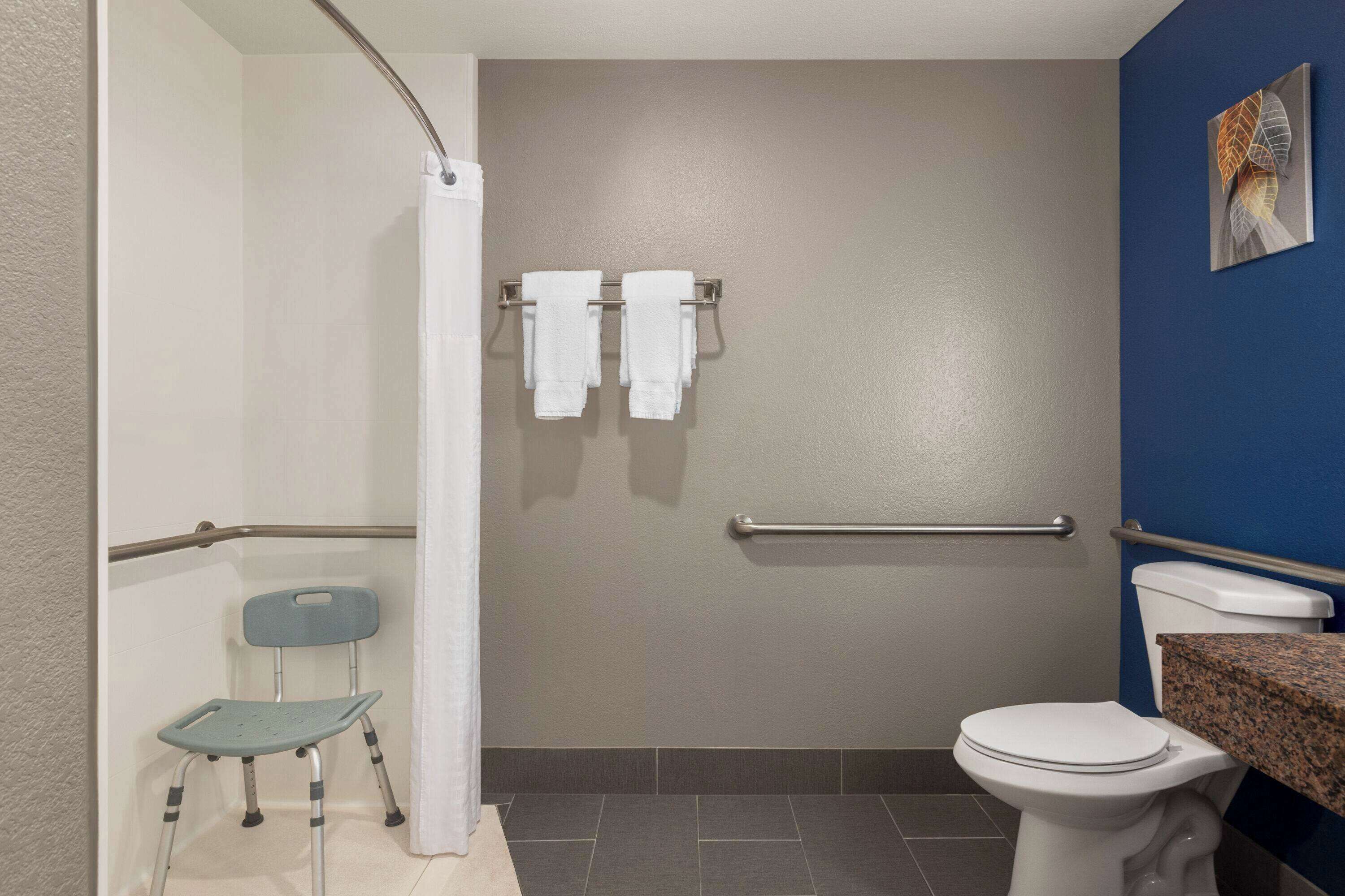 A bathroom with a shower, a toilet, a grab bar, a chair, and a towel rack.