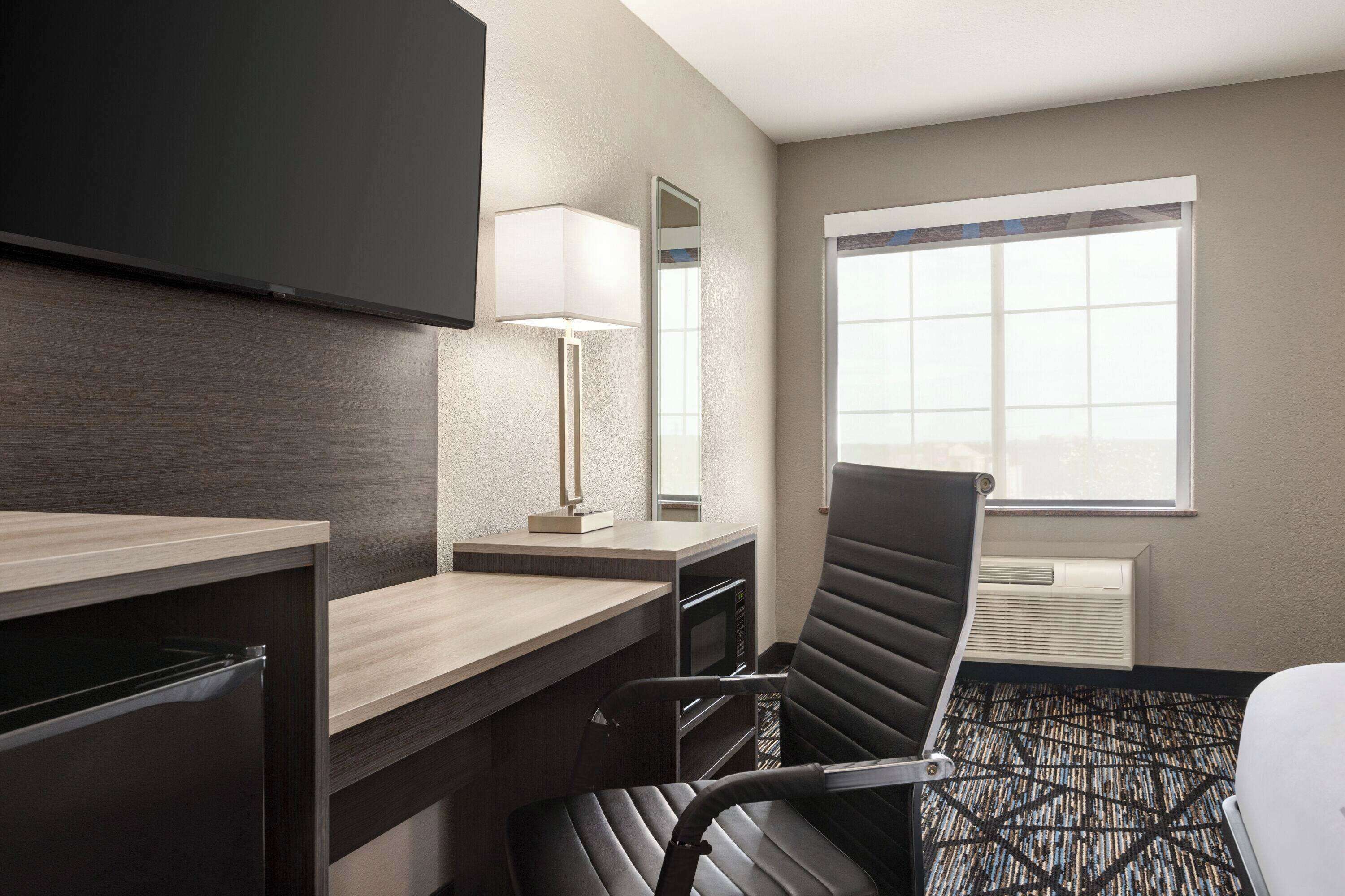 A hotel room with a desk, chair, flat-screen TV, lamp, and a large window.