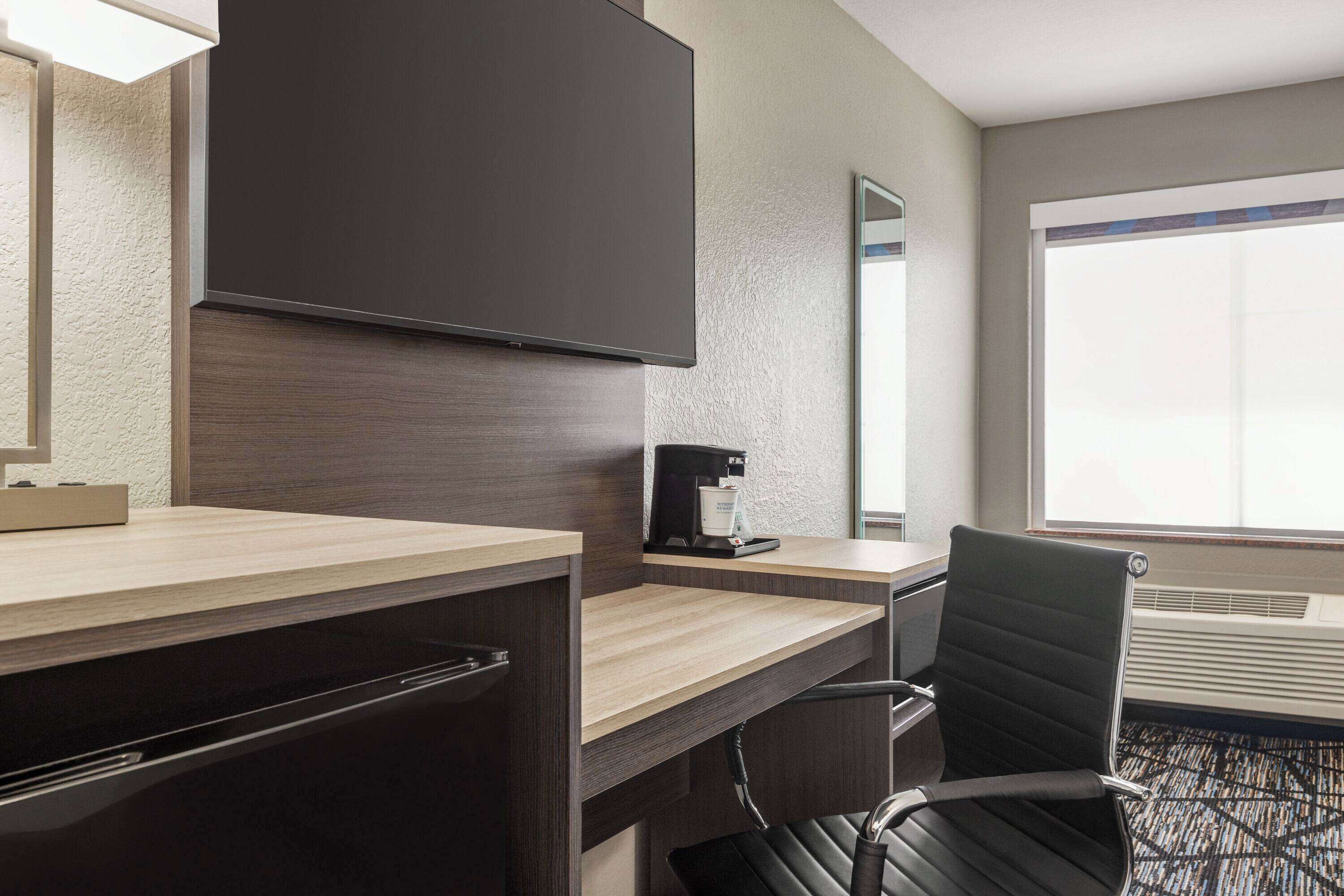 A hotel room with a wooden desk, a black leather chair, a flat-screen TV mounted on the wall, a coffee maker, and a large window providing natural light.