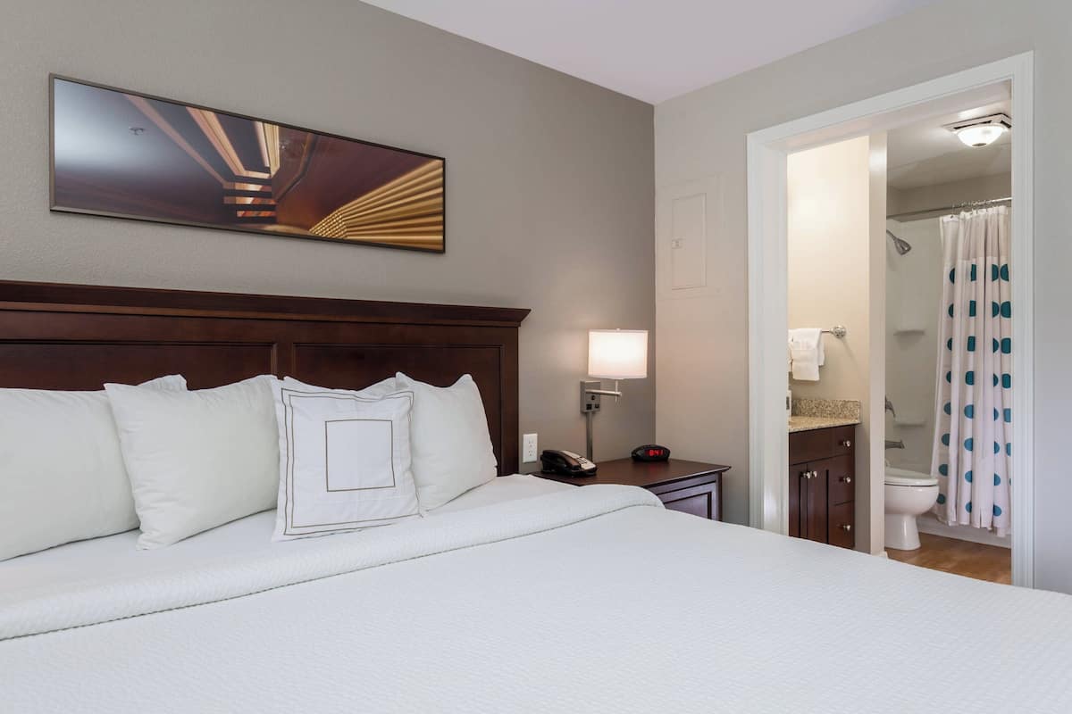 A hotel room with a large bed, a bedside table with a lamp, a telephone, and a bathroom visible through an open door.
