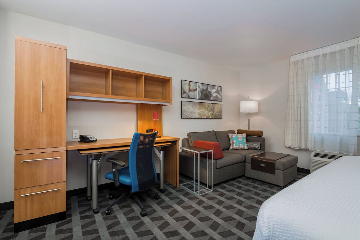 A hotel room with a wooden desk, a blue office chair, a sofa, a bed, and a window with curtains.