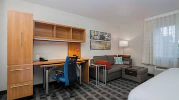 A hotel room with a wooden desk, a blue office chair, a sofa, a bed, and a window with curtains.