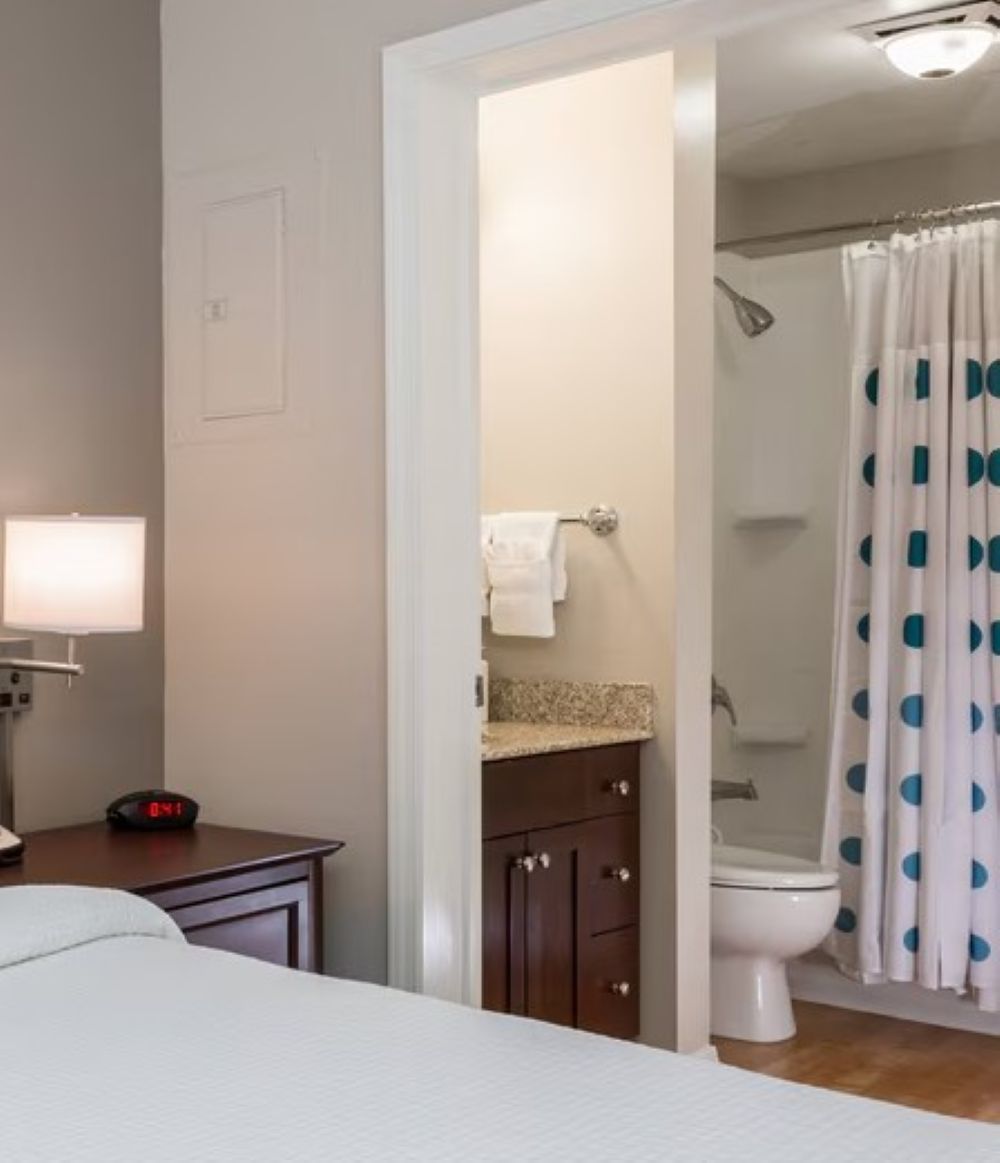 Een hotelkamer met een bed, een nachtkastje met een lamp, een digitale klok, een badkamer met toilet en douche, en een gordijn met stippen.