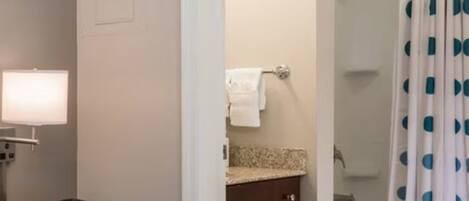 Een hotelkamer met een bed, een nachtkastje met een lamp, een digitale klok, een badkamer met toilet en douche, en een gordijn met stippen.