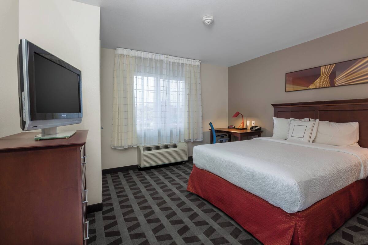 A hotel room with a large bed, a desk with a chair, a flat-screen TV, and a window with curtains.