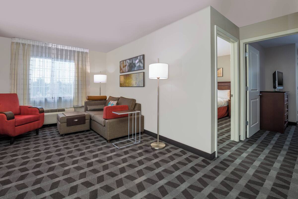 A hotel room with a sofa, armchair, and a small table. There is a lamp, a TV, and a wardrobe.