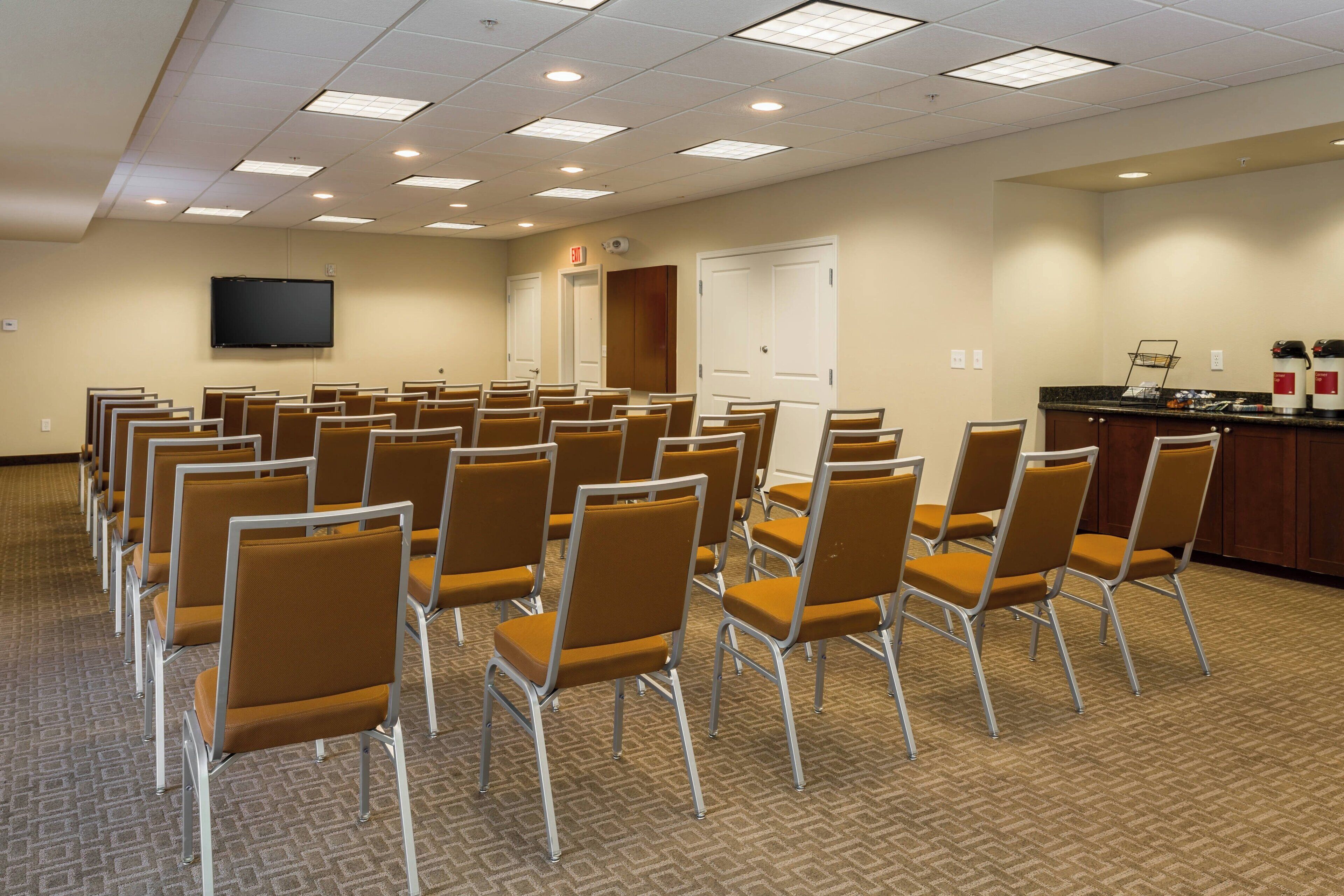 A conference room with rows of chairs, a flat-screen TV, a door, and a counter with a coffee machine.