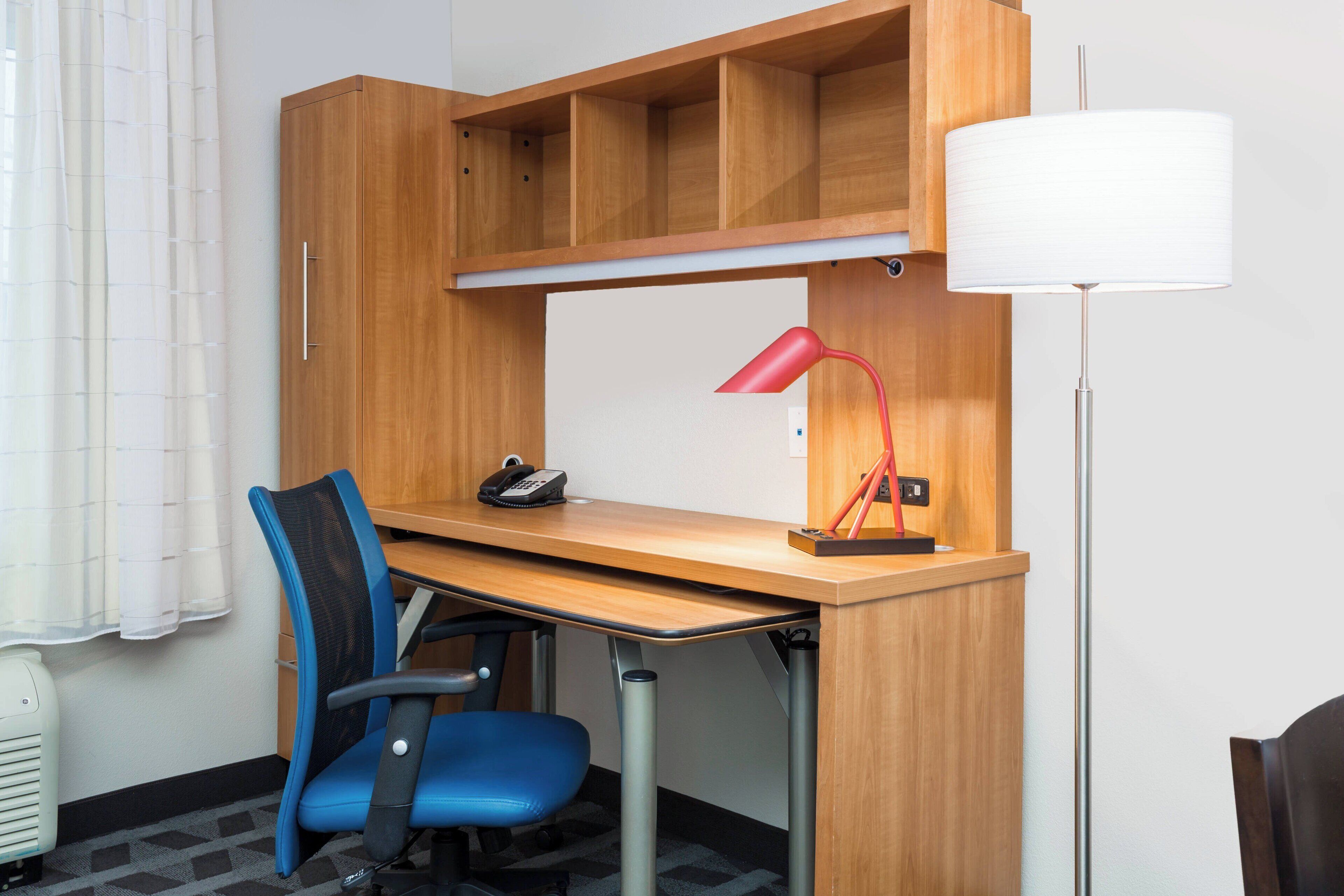 Quarto de hotel com escrivaninha de madeira, cadeira de escritório azul, abajur vermelho e luminária de chão.