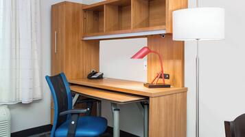 Quarto de hotel com escrivaninha de madeira, cadeira de escritório azul, abajur vermelho e luminária de chão.