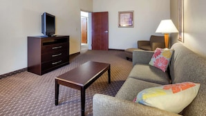 Habitación de hotel con un sofá, un televisor sobre un mueble de madera, una lámpara y una puerta que da a otra habitación.