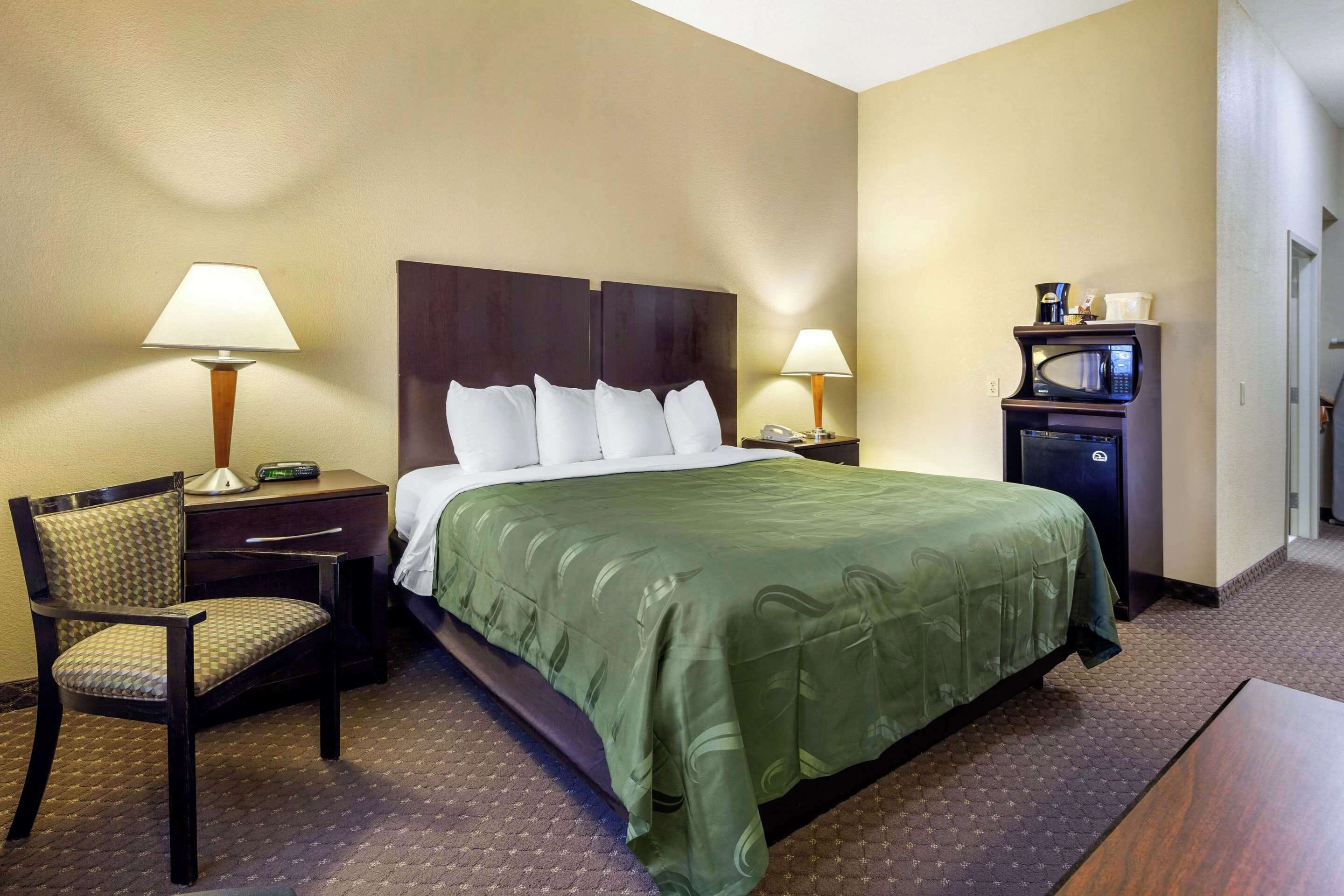A hotel room with a bed, two bedside tables, a desk with a chair, a microwave, and a small refrigerator.