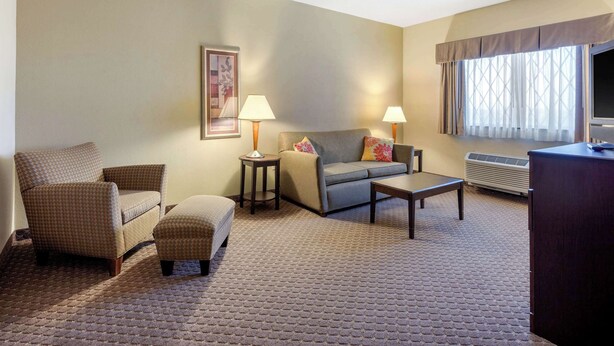 Habitación de hotel con sofá, sillón, reposapiés, dos mesitas de noche con lámparas, una mesa de centro, televisor y ventana con cortinas.