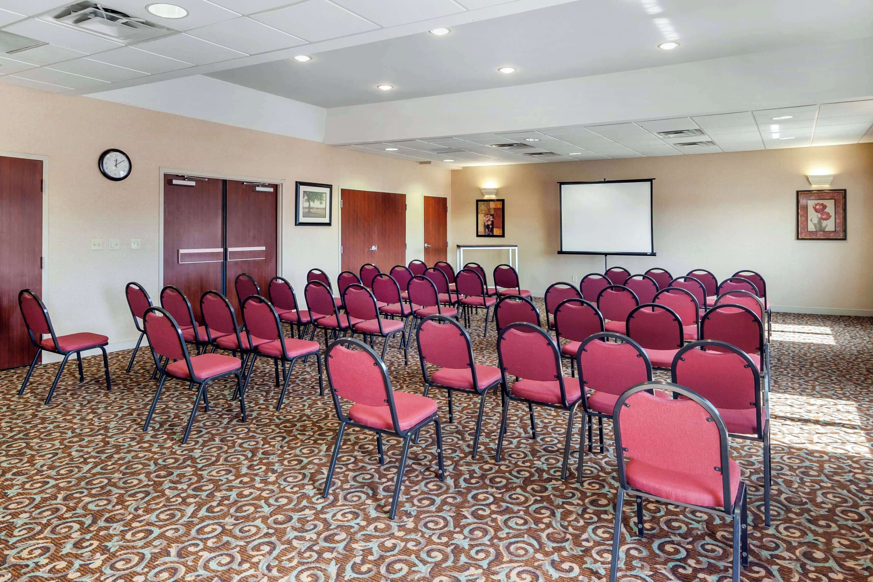 Une salle de conférence avec des rangées de chaises rouges, un écran de projection et une horloge au mur.