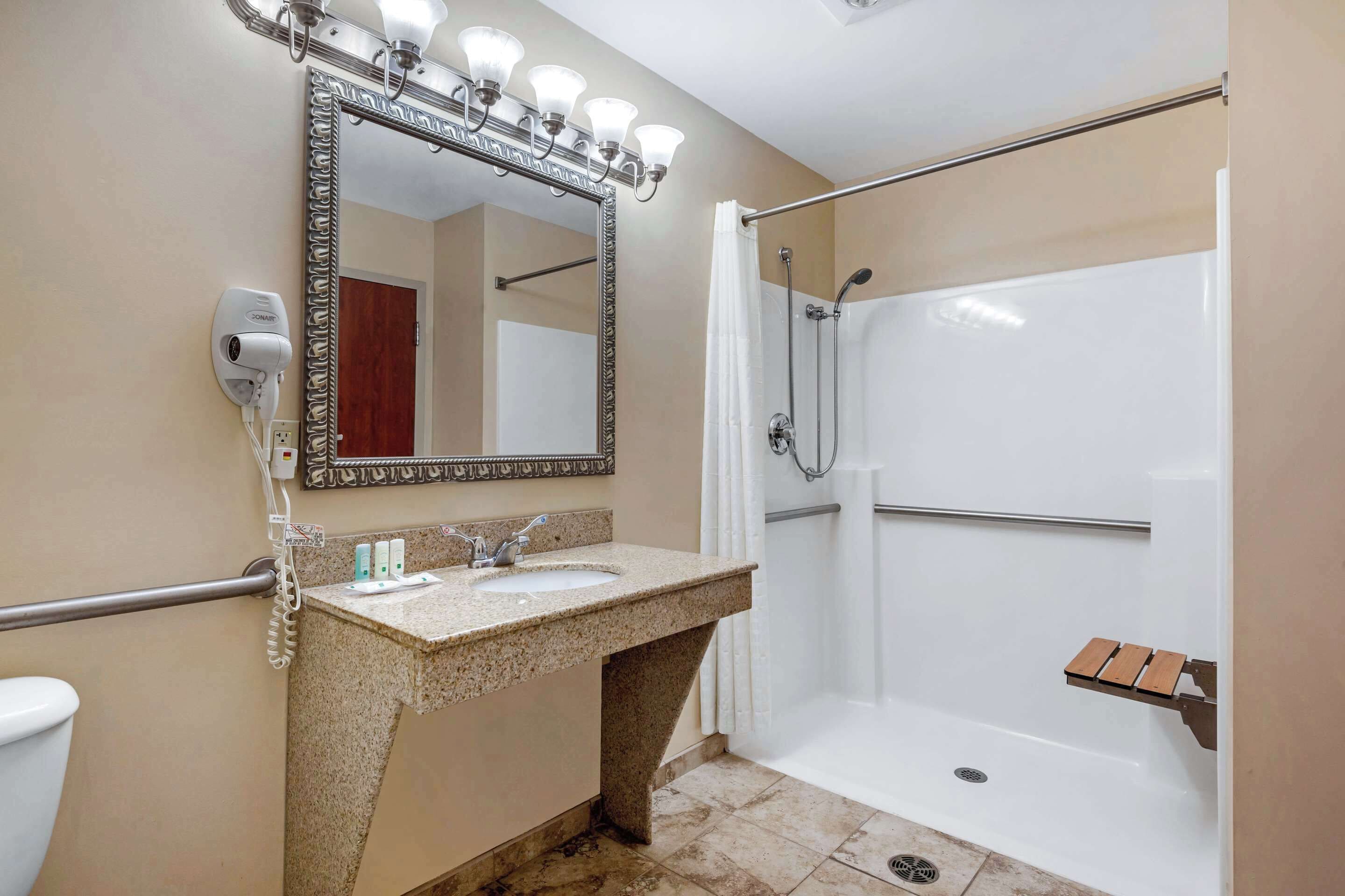 A bathroom with a granite sink, a large mirror, a hairdryer, a shower, and a wooden bench.