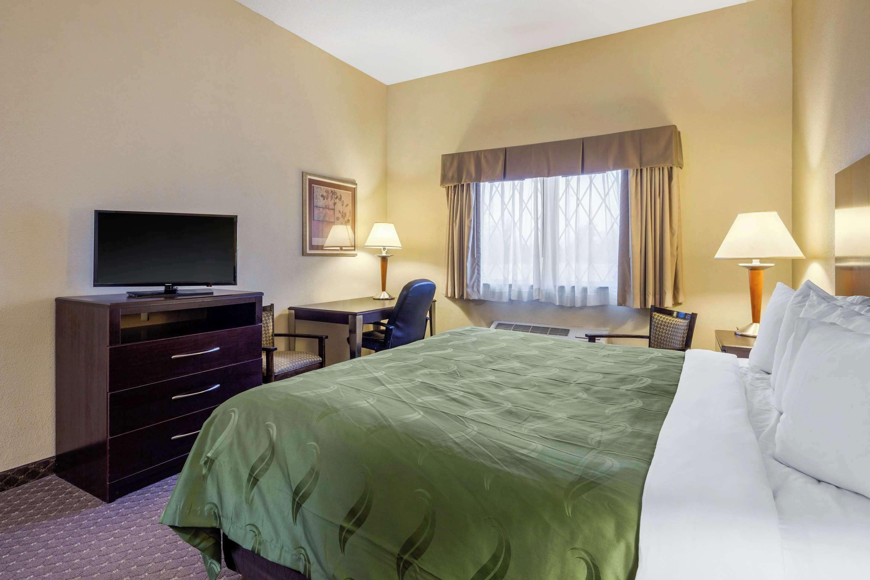 Habitación de hotel con cama, escritorio, silla, televisor y ventana con cortinas.