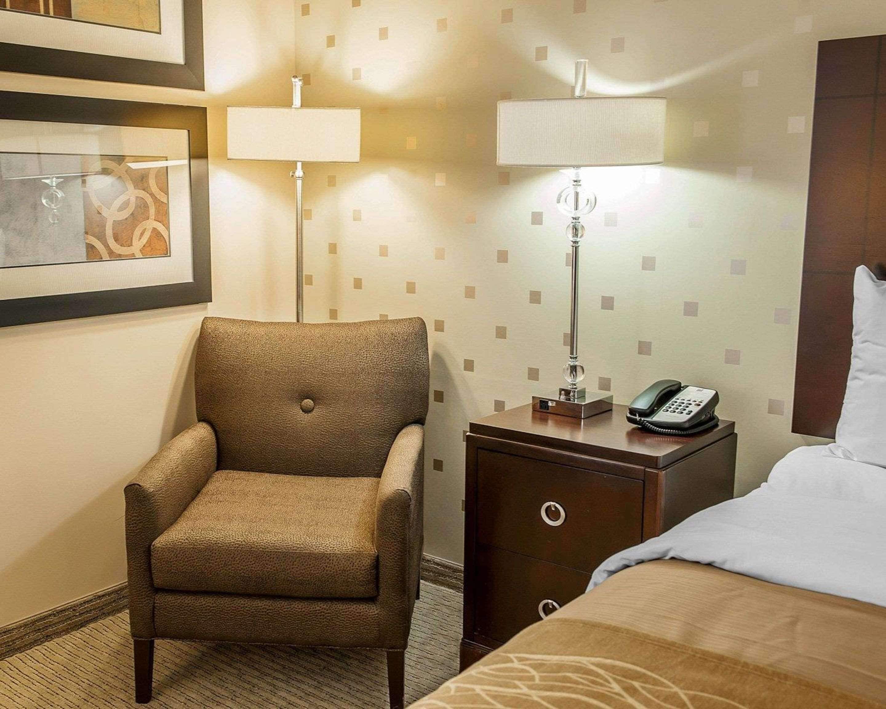 A hotel room with a single brown armchair, a bedside table with a lamp, a phone, and a bed with a white pillow.