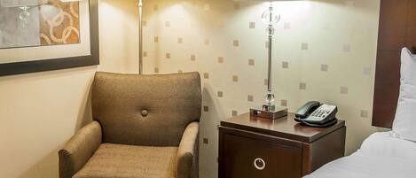 A hotel room with a single brown armchair, a bedside table with a lamp, a phone, and a bed with a white pillow.