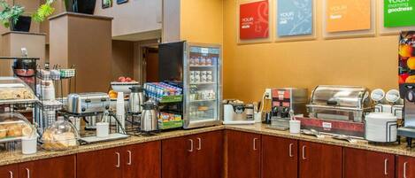 A hotel breakfast area with a self-service station, refrigerated display, and various food items.
