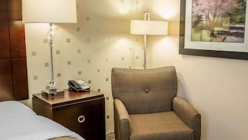 A hotel room with a bed, a nightstand with a lamp and phone, a leather armchair, and a framed picture on the wall.