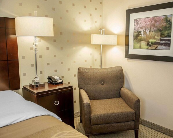 A hotel room with a bed, a nightstand with a lamp and phone, a leather armchair, and a framed picture on the wall.