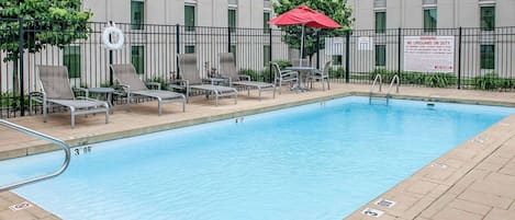 A swimming pool with lounge chairs and a red umbrella, surrounded by a fence and a building with multiple windows.