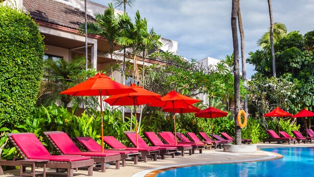A poolside area with red umbrellas and lounge chairs, surrounded by lush greenery and a building with a tiled roof. - Coconut Village Resort (Patong)