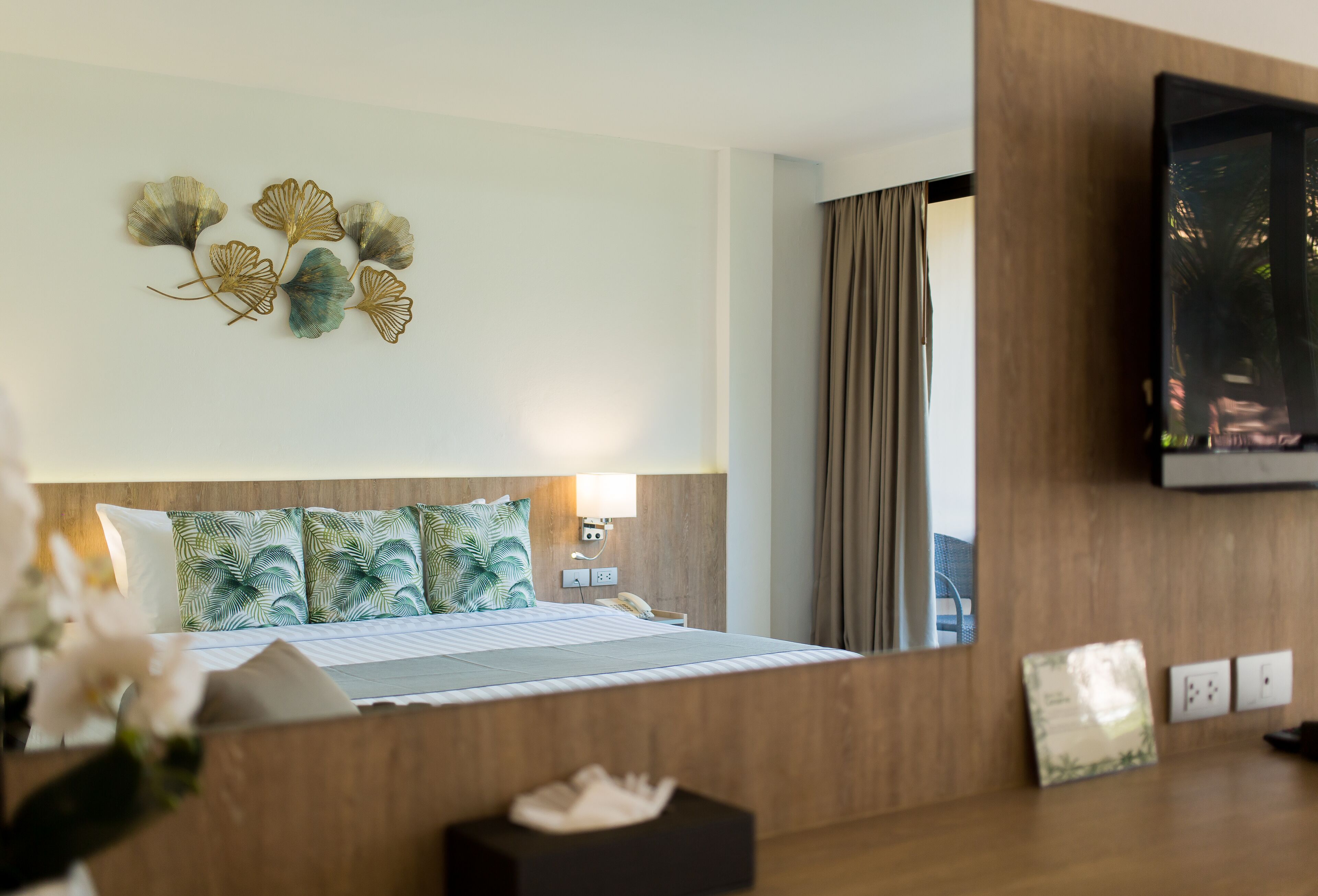A hotel room with a bed, a wall-mounted TV, and a decorative leaf art piece.
