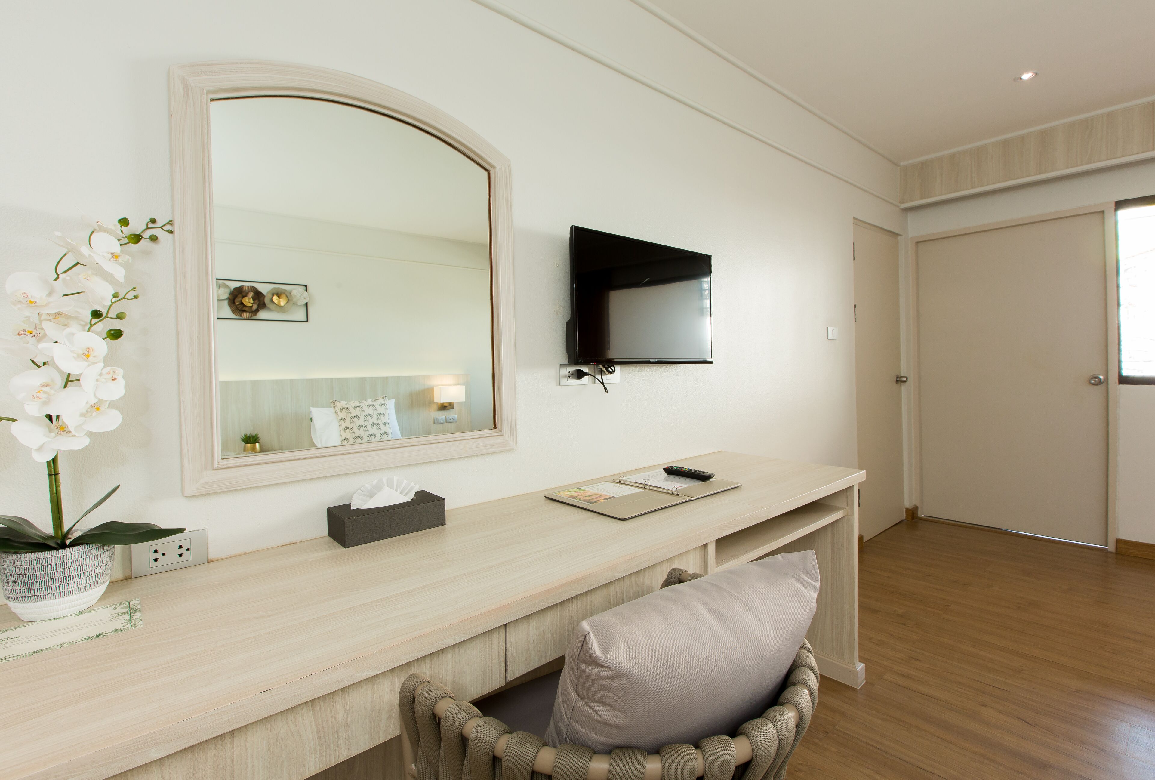 A hotel room with a large mirror, a flat-screen TV mounted on the wall, a wooden console table, a wicker chair, and a door leading to another room.