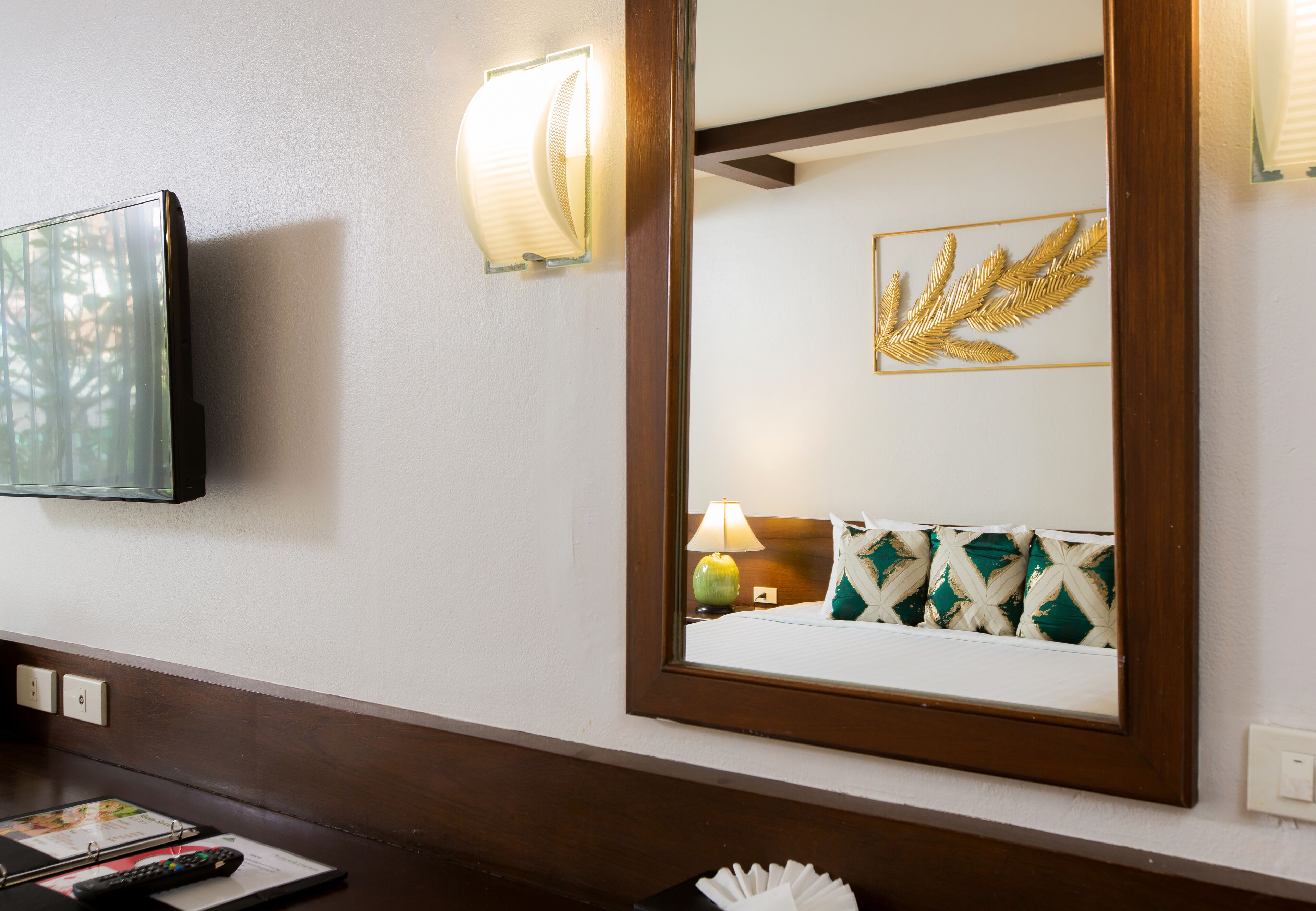 A hotel room with a large mirror, a television mounted on the wall, a bedside table with a lamp, and a decorative wall piece.